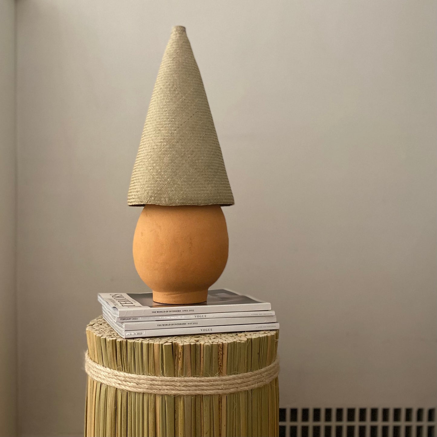 The Duende Lamp has a handcrafted, round, terracotta colored, ceramic base, with a natural colored, pointed cone shaped, hand woven, dried palm leaf lampshade.