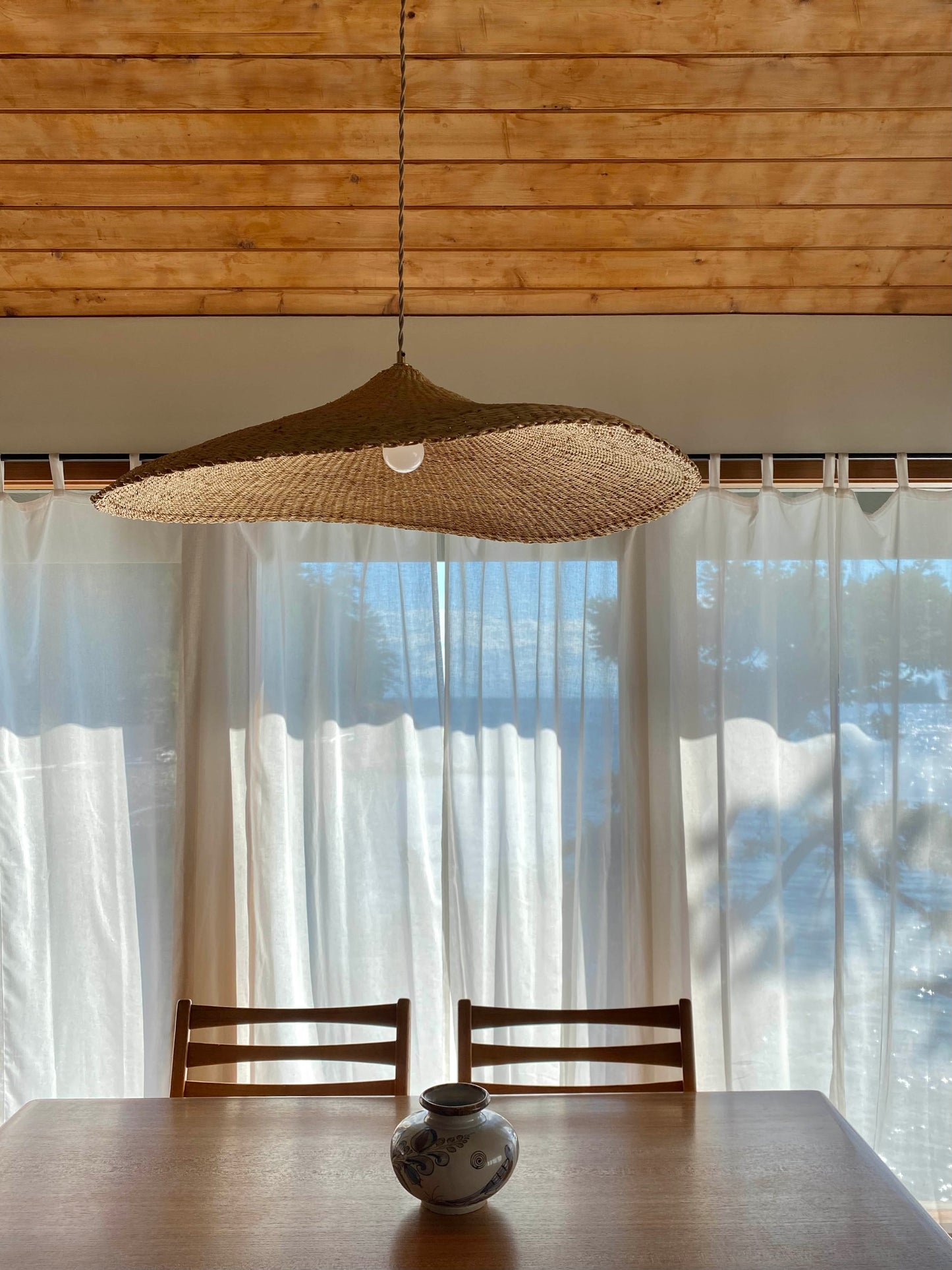 The Sunhat pendant light is a shallow cone shape, handwoven from natural colored, sustainable elephant grass in northern Ghana. With its classic shape this pendant is ideal for any living space. Show here over a dining table.