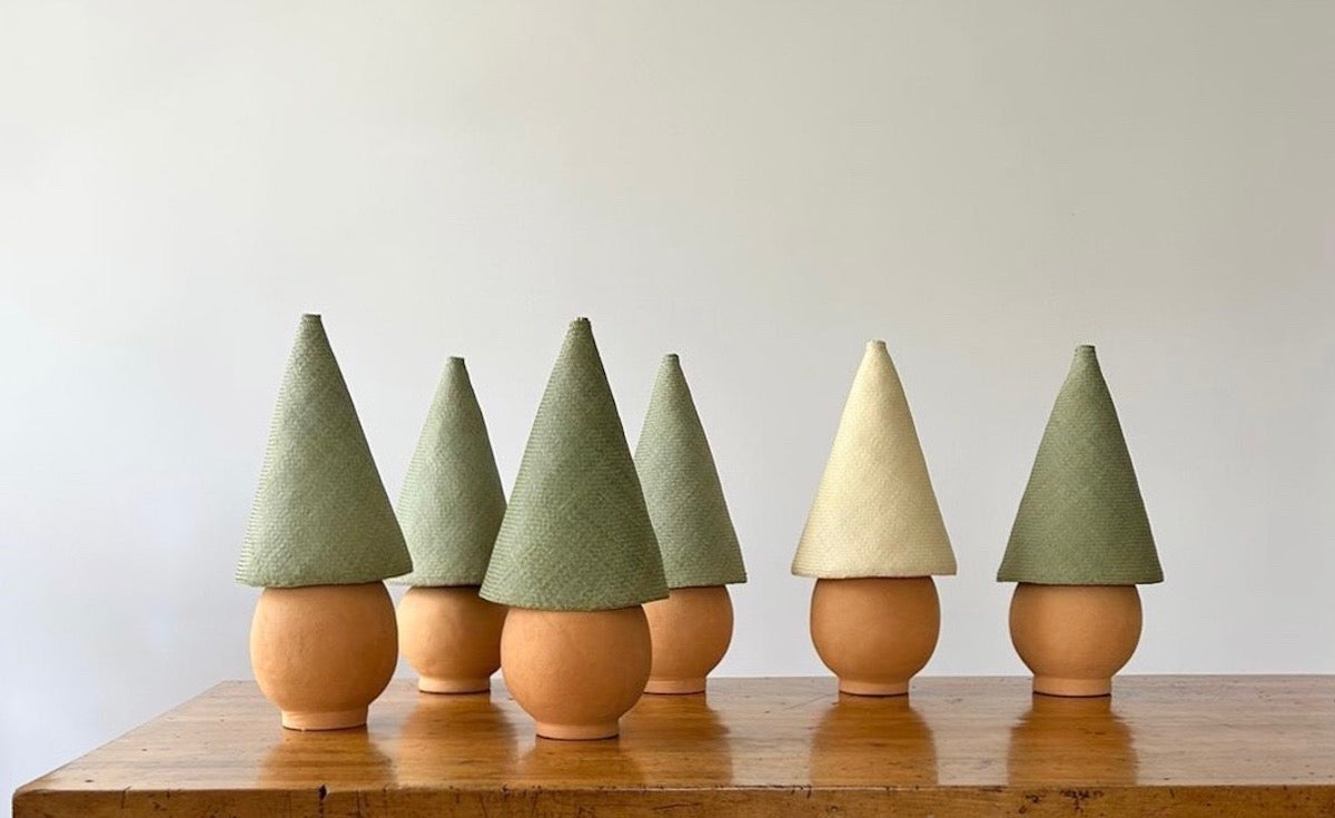 Shown are six Duende lamps together on a table. The Duende Lamp has a handcrafted, round, terracotta colored, ceramic base, with a natural colored, pointed cone shaped, hand woven, dried palm leaf lampshade.