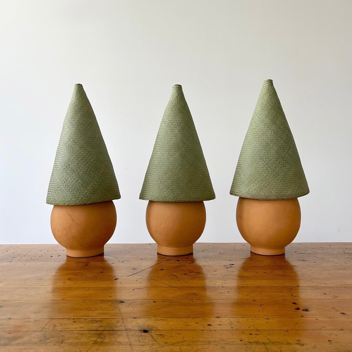 The Duende Lamp has a handcrafted, round, terracotta colored, ceramic base, with a natural colored, pointed cone shaped, hand woven, dried palm leaf lampshade. Here are three of the lamps shown together