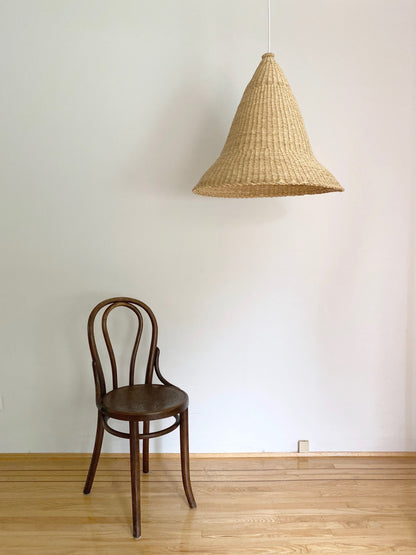 Witches Hat light shade shown with a chair. Shade is handwoven. Their steep cone shape casts a downward light, ideal for spaces that call for a more direct light source.